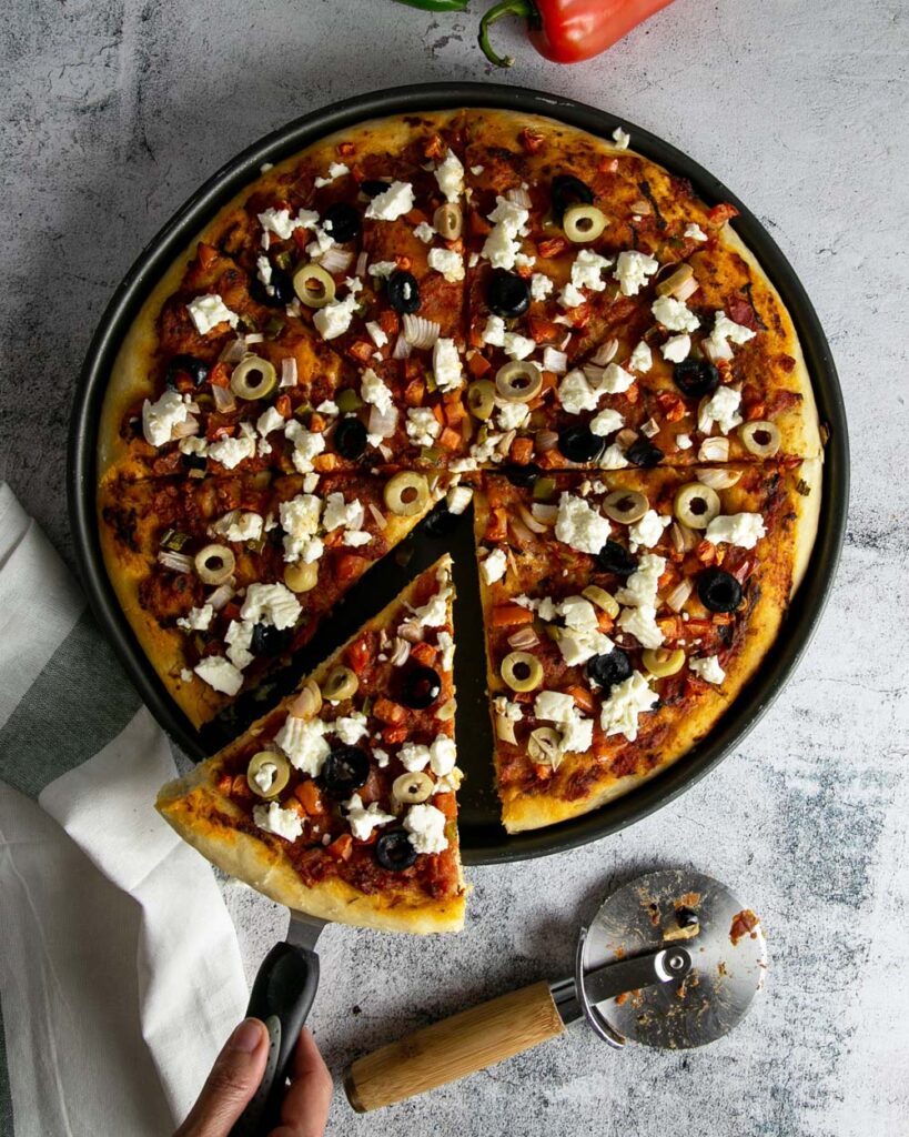 A sliced up Greek style pizza on a pizza pan with a white marble background.