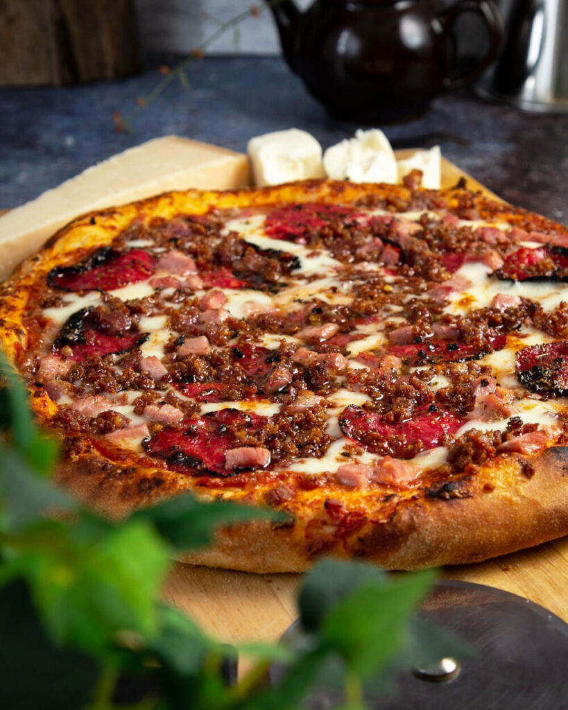 Side view of meat lovers pizza on a wood cutting board behind some fresh basil.