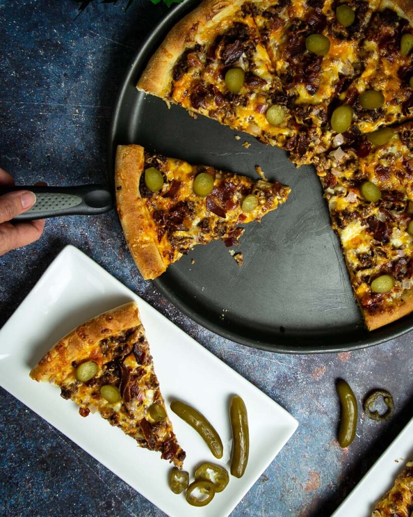 Serving cheeseburger pizza onto a while plate with jalapeños.