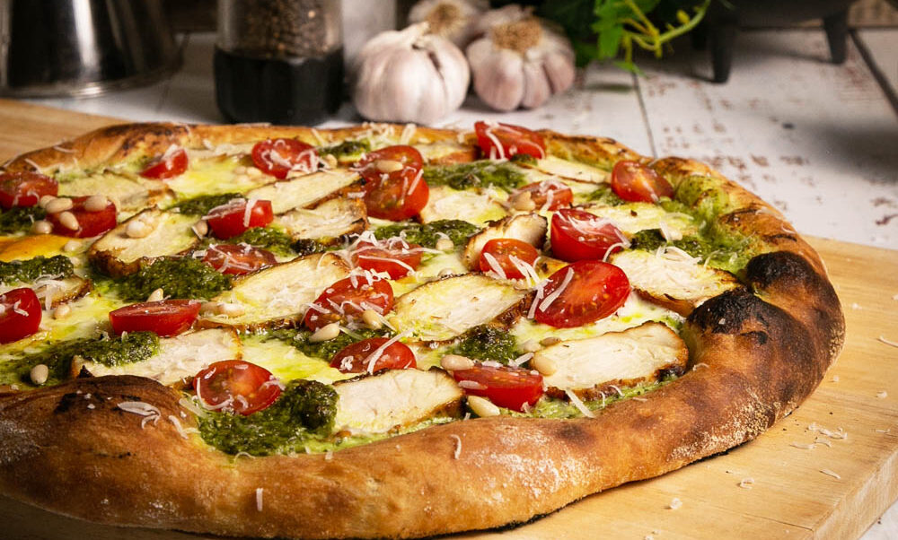 Side view of chicken pesto pizza on a cutting board with vegetables in the background