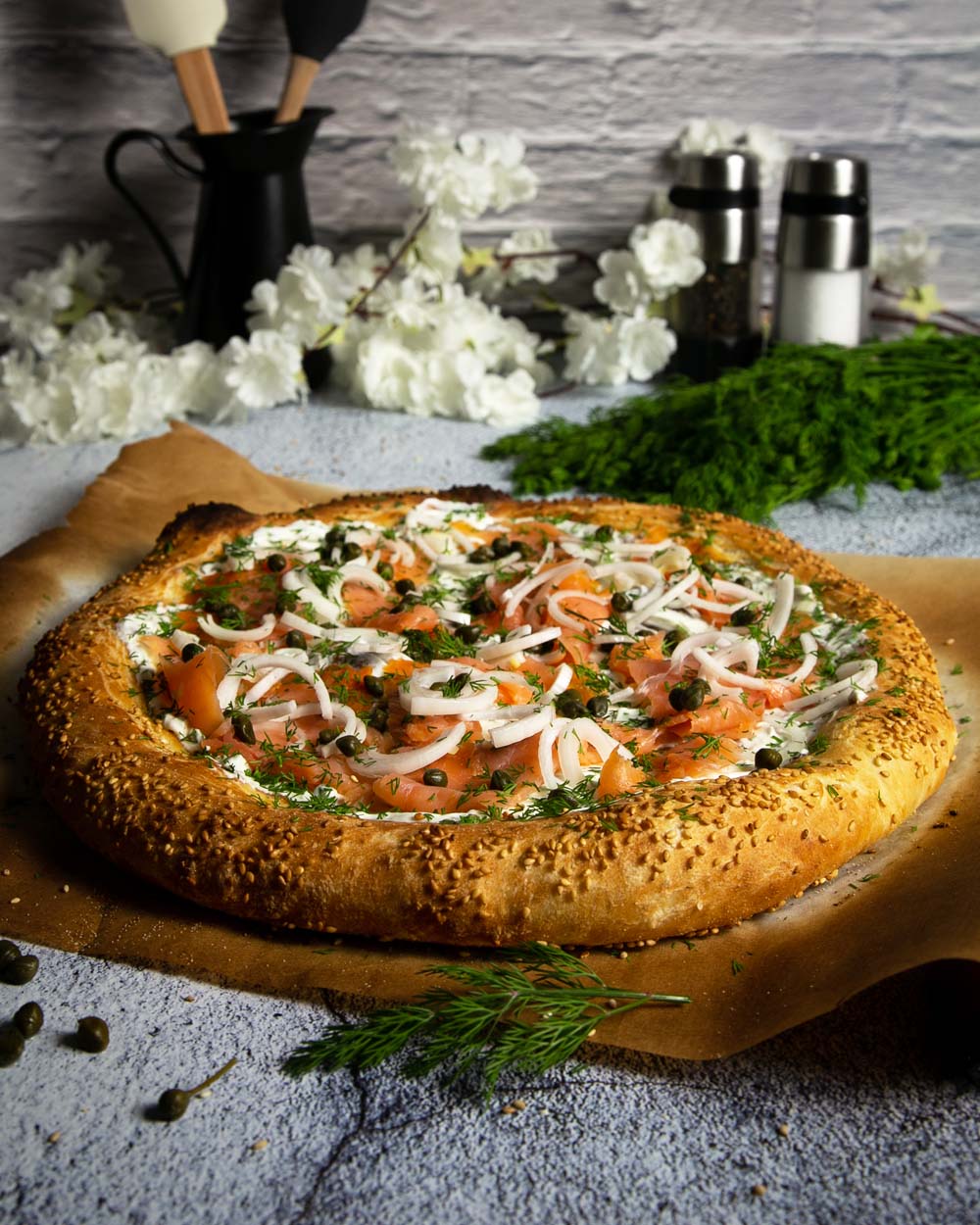 Side view of deli style breakfast pizza on a white granite counter with flowers and fresh herbs 