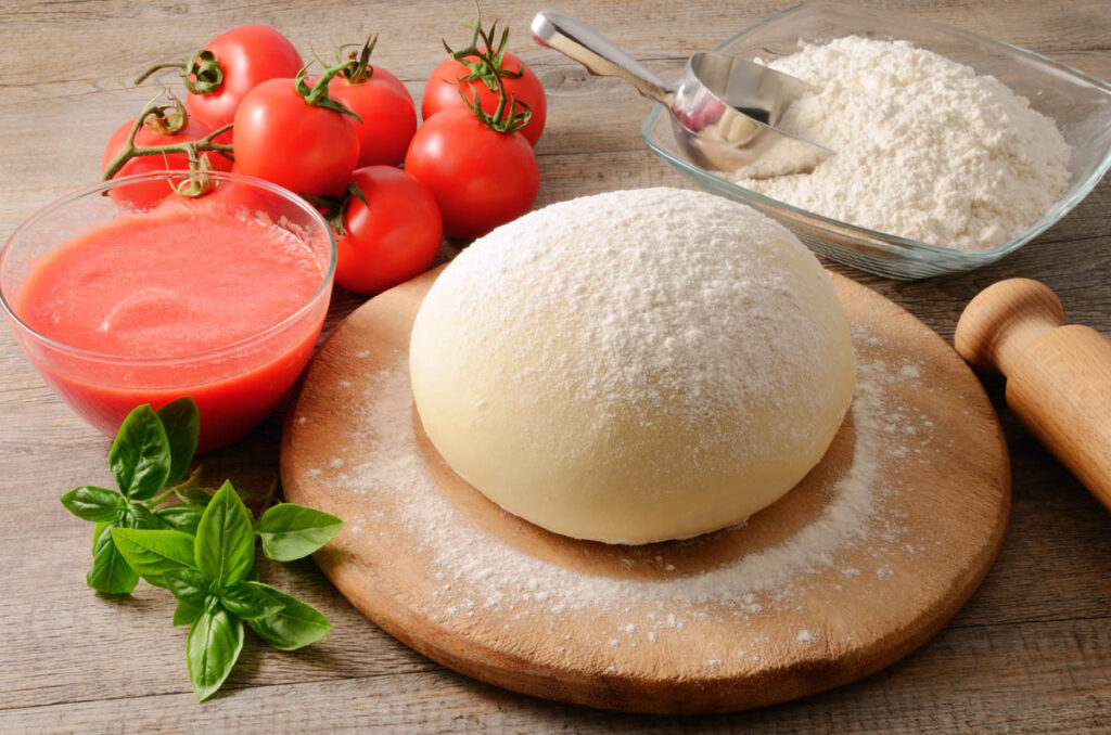 Proofed pizza dough for the best pizza crust on a round cutting board surrounded by pizza toppings