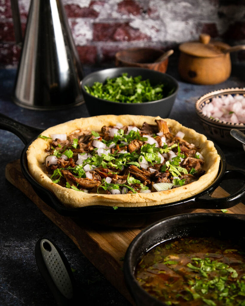 side view of birria pizza with a side of consume on a wood and marble background